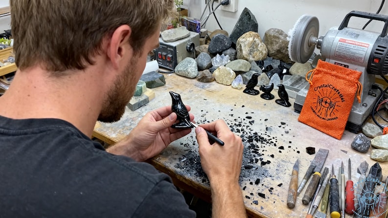 May include: A person working on a black bird figurine with a tool. Several other bird figurines are on the table, along with an orange bag that says "crystalcrafthel". A grinding wheel and various stones are in the background.