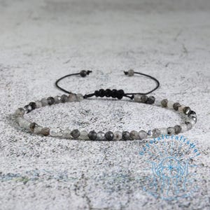 May include: A delicate bracelet featuring small, faceted beads in shades of gray and clear. The beads are strung on a black cord with an adjustable sliding knot closure. The bracelet is designed for a minimalist aesthetic.