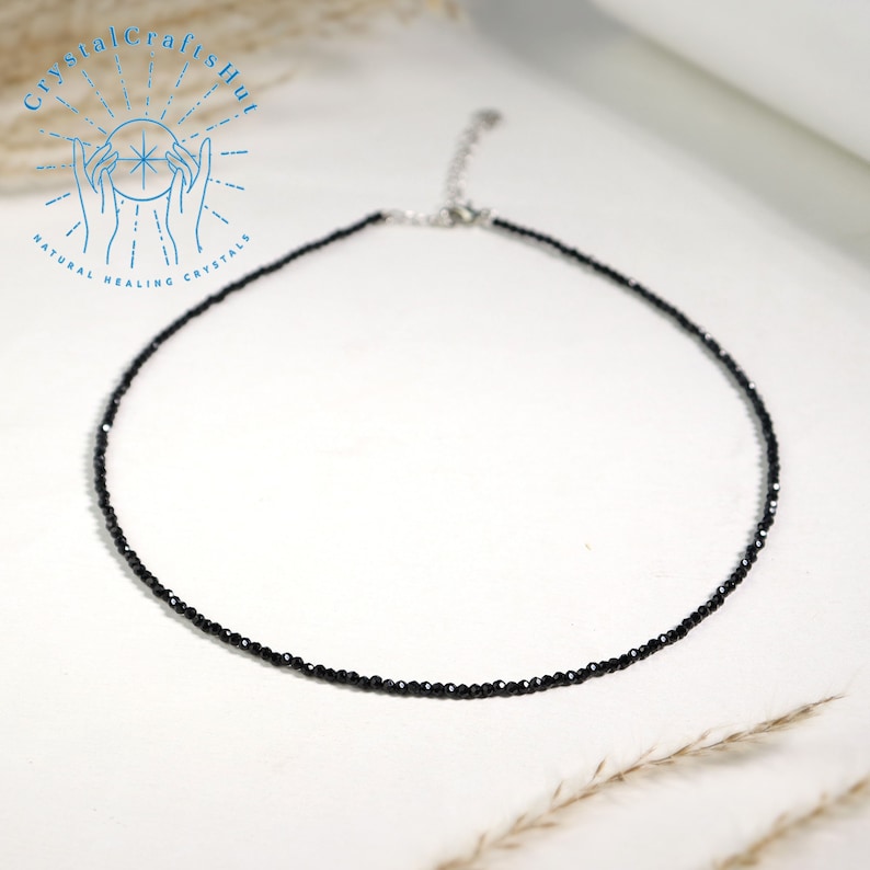 K&ouml;nnte beinhalten: Eine zarte schwarze Choker-Kette aus kleinen schwarzen Edelsteinperlen. Die Kette liegt auf einem wei&szlig;en Hintergrund.