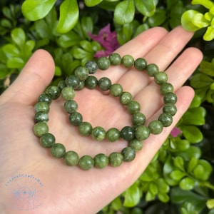 May include: Two green jade stone bracelets with round beads. The bracelets are stacked on top of each other and are being held in a hand.