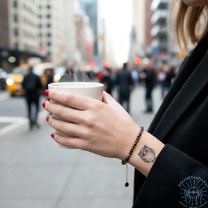 May include: A person holding a white ceramic mug with steam rising from it. A beaded bracelet with a gold chain and a small owl tattoo are visible on the wrist. The background shows a city street with blurred figures.