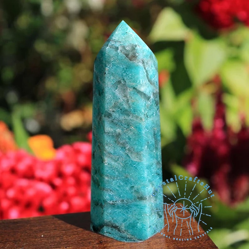 May include: A tall, turquoise Amazonite crystal obelisk with a pointed top. The stone has a marbled appearance with dark gray veining. The obelisk is displayed on a wooden surface, with a blurred background of red and green foliage.