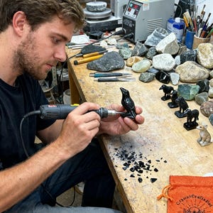 May include: A person using a rotary tool to carve a small, black raven figurine. The wooden workbench is covered with tools, gemstones, and other carved animal figurines. An orange bag with text is on the right.