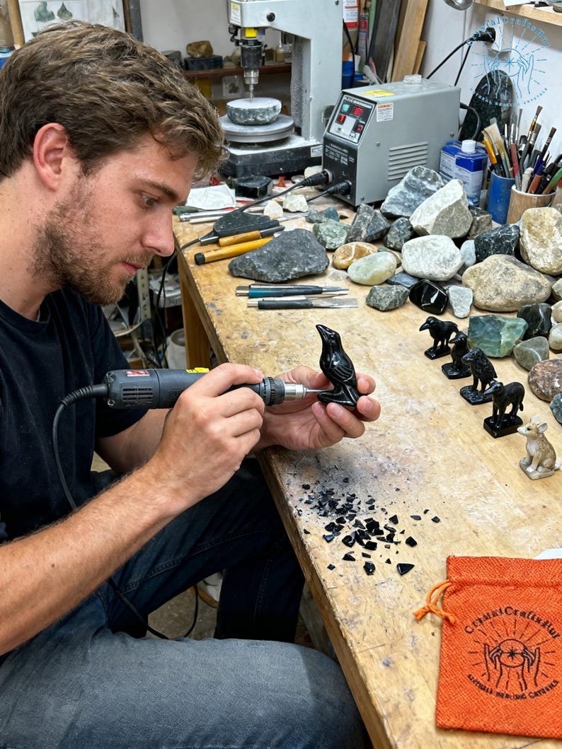 May include: A person using a rotary tool to carve a small, black raven figurine. The wooden workbench is covered with tools, gemstones, and other carved animal figurines. An orange bag with text is on the right.