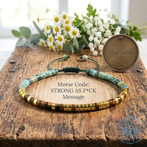 May include: A beaded bracelet with turquoise and gold-colored beads, and brown tiger eye beads. The bracelet is on a wooden surface with flowers and a metal disc that says "CrystalCraftsHut". The text on the image says "Morse Code: STRONG AS F*CK Message".