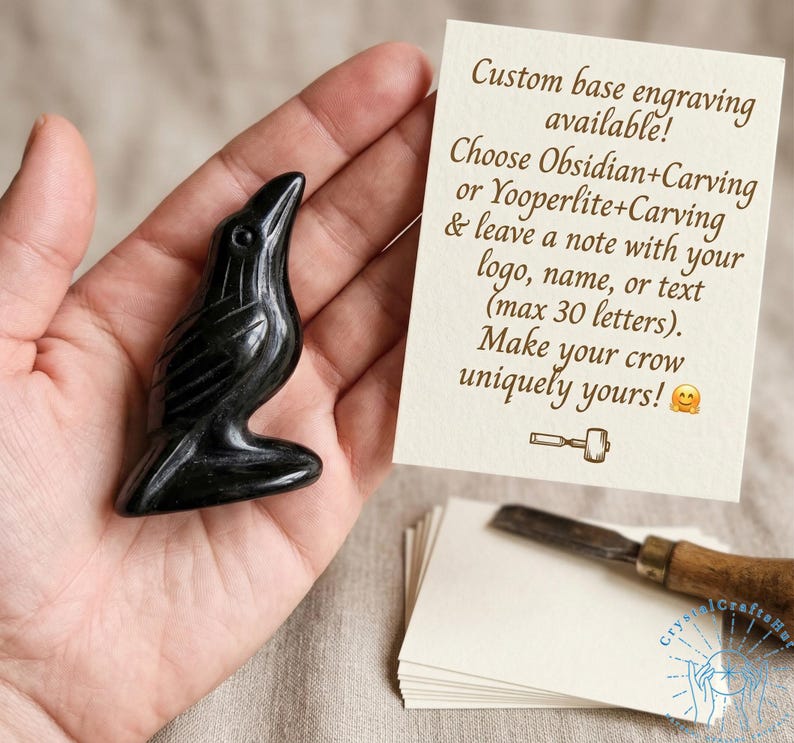 May include: A black obsidian crow figurine with a glossy finish. The crow is perched on a small base. A card in the background states "Custom base engraving available!" and "Make your crow uniquely yours!"