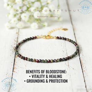 May include: A delicate bracelet featuring small, round bloodstone beads in shades of green and burgundy, strung on a gold-colored chain. The bracelet is displayed on a white wooden surface, with text that reads "Benefits of Bloodstone: Vitality & Healing, Grounding & Protection."