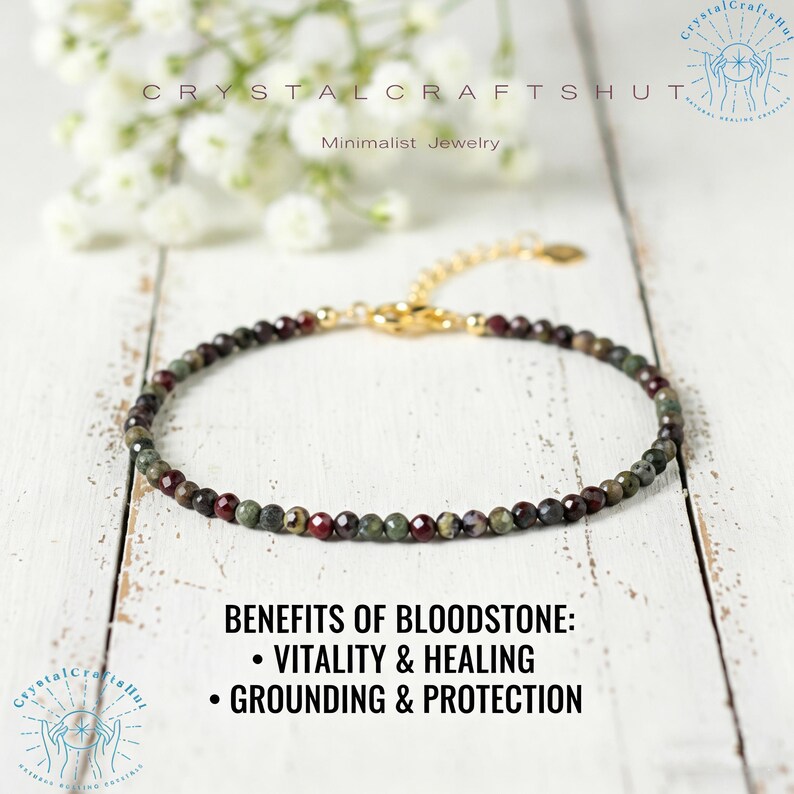 May include: A delicate bracelet featuring small, round bloodstone beads in shades of green and burgundy, strung on a gold-colored chain. The bracelet is displayed on a white wooden surface, with text that reads "Benefits of Bloodstone: Vitality & Healing, Grounding & Protection."