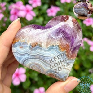 May include: A heart-shaped stone sculpture with a mix of purple, brown, and blue hues. The stone has a layered, agate-like appearance. The words "KEEP GOING 2024" are engraved on the front. The sculpture is held in a hand.