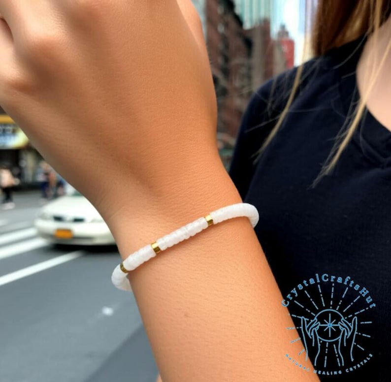 May include: White beaded bracelet with gold-colored accents. The bracelet is worn on a wrist, showcasing its delicate design. The beads are small and round, creating a minimalist aesthetic. The bracelet is a piece of jewelry.