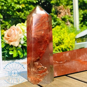 May include: A clear quartz crystal point with red and brown inclusions. The crystal is approximately 5 inches tall and has a pointed tip. The crystal is sitting on a wooden surface.