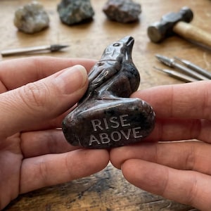 May include: A carved stone raven figurine with the words "RISE ABOVE" etched on the base. The raven is dark gray and brown. The figurine is held in a person's hands. Other stones and tools are in the background.