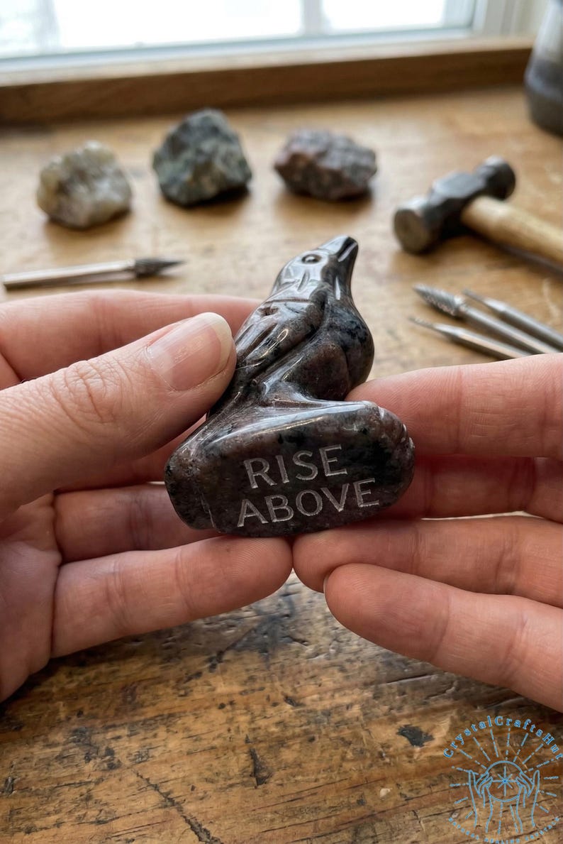 May include: A carved stone raven figurine with the words "RISE ABOVE" etched on the base. The raven is dark gray and brown. The figurine is held in a person's hands. Other stones and tools are in the background.