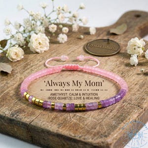 Peut inclure: Un bracelet de perles roses et violettes avec des détails dorés. Le bracelet porte l'inscription "Always My Mom" et est composé d'améthyste et de quartz rose. Il est présenté sur une planche de bois avec des fleurs blanches.