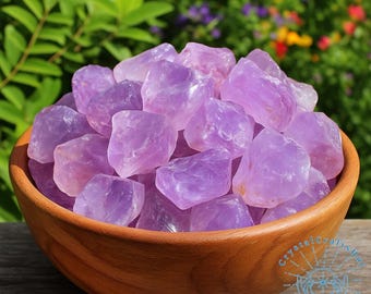 May include: A wooden bowl filled with raw, light purple amethyst crystals. The crystals have a natural, rough texture and varying shades of lavender. The bowl is made of polished wood, and the background features green foliage and colorful flowers.