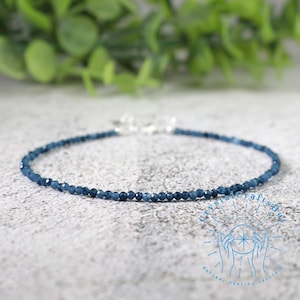 May include: A delicate blue beaded bracelet with a silver clasp. The bracelet is made of small, round, dark blue beads. The background is a light gray surface with green foliage.