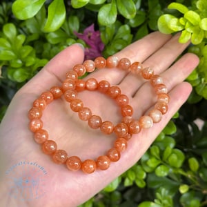 May include: Two orange sunstone bead bracelets. The bracelets are made of round, polished beads with a shimmering, reflective quality. The bracelets are displayed against a backdrop of green foliage.