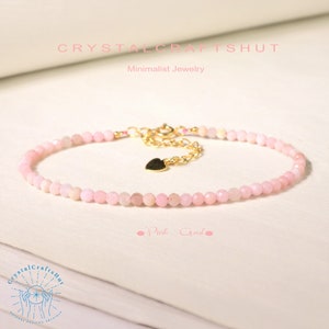May include: A delicate pink opal beaded bracelet with a gold heart charm and a gold chain closure. The bracelet is on a white surface with the text "CrystalCraftsHut" and "Minimalist Jewelry".