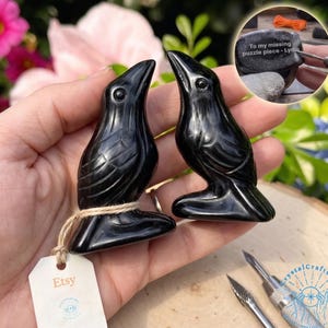 May include: Two glossy black raven figurines with detailed feather carvings. Each raven is perched on a small base and tied with twine. The Etsy tag is visible. A small stone with text is in the background.