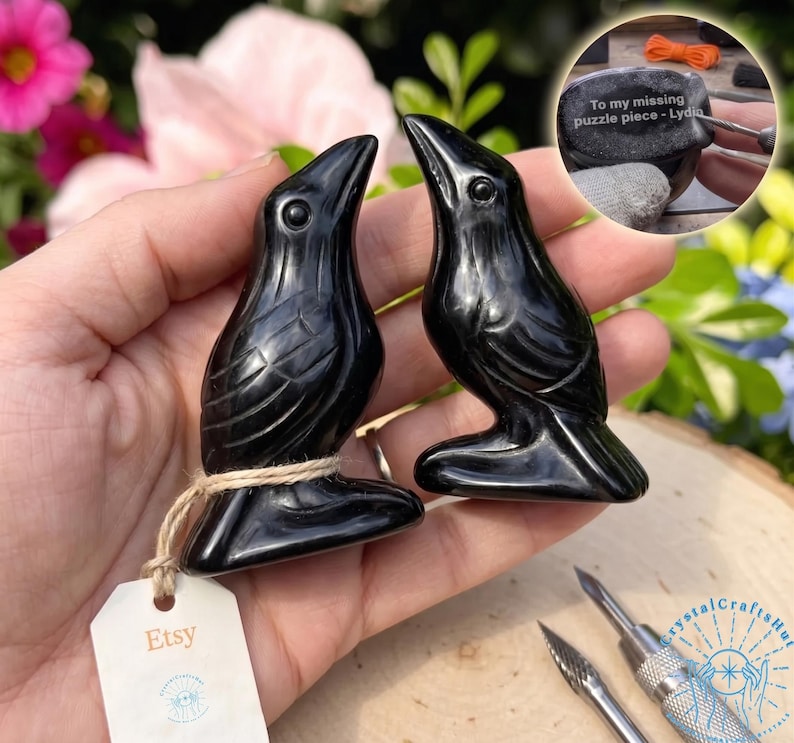 May include: Two glossy black raven figurines with detailed feather carvings. Each raven is perched on a small base and tied with twine. The Etsy tag is visible. A small stone with text is in the background.