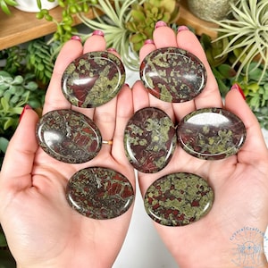 May include: Seven oval-shaped stones with a mix of red and green patterns are displayed. The stones are smooth and polished, with a natural, earthy appearance. The stones are held in open hands, showcasing their size and shape.