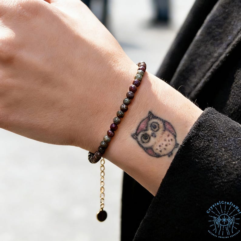 May include: A beaded bracelet with small, round, dark red and green stones. The bracelet has a gold-colored chain and a small, round charm. The bracelet is worn on a wrist with an owl tattoo.