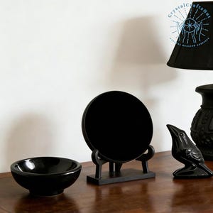 May include: A collection of black decorative items on a wooden surface. The items include a round mirror on a stand, a small bowl, and a bird figurine. A black lamp and vase are also visible in the background.