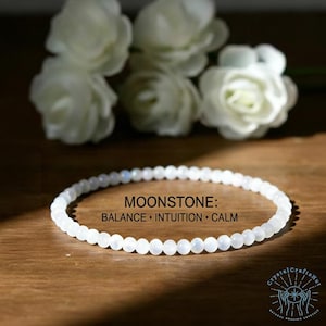 May include: A delicate white moonstone bead bracelet. The bracelet is strung with small, round, polished beads. Text on the image reads "MOONSTONE: BALANCE • INTUITION • CALM". White flowers are in the background.