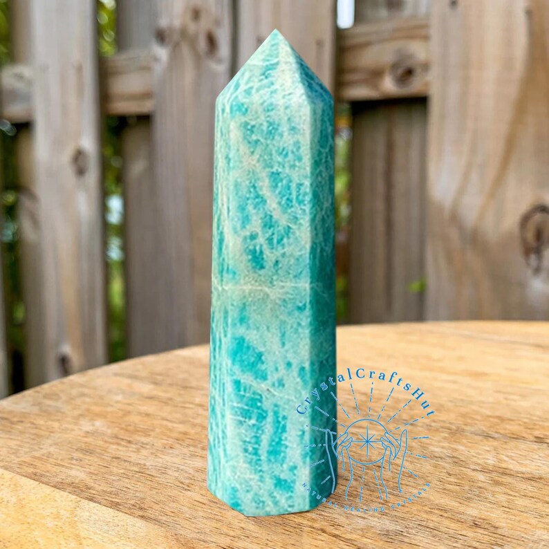 May include: A tall, turquoise amazonite crystal obelisk with white veining. The polished stone has a faceted top and a rectangular base. The obelisk is displayed on a wooden surface with a blurred background of a wooden fence and greenery.