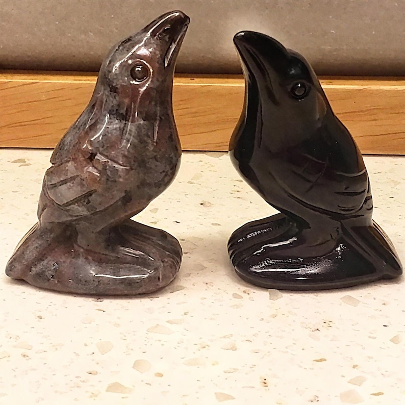 May include: Two carved stone raven figurines. One is a mottled gray and brown, the other is a glossy black. Both are perched with their heads tilted upwards, showcasing detailed craftsmanship and a polished finish. The ravens are displayed on a speckled surface.