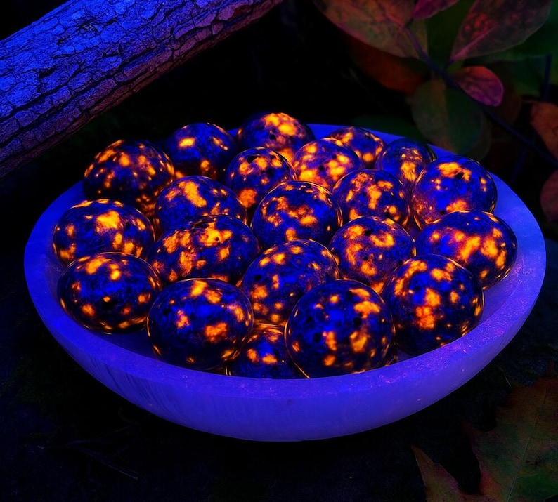 May include: A collection of glowing, spherical stones with a blue and orange speckled pattern, displayed in a light blue bowl. The stones are illuminated, creating a vibrant and eye-catching display. The bowl is set against a dark background with hints of foliage.