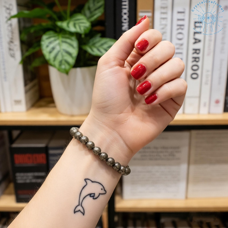 May include: A pyrite beaded bracelet is worn on a wrist with a dolphin tattoo. The hand has red nail polish. The background includes a bookshelf with books and a potted plant.