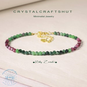 Op de afbeelding: Een delicate armband gemaakt met groene zoisiet en robijn kralen. De armband heeft een gouden sluiting en een klein hartje bedel. De armband ligt op een wit oppervlak met de tekst "Ruby Zoisite" en "CrystalCraftsHut" eronder.