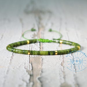 May include: A green jade gemstone bracelet with gold accents and a green string closure. The bracelet is on a white wooden surface.