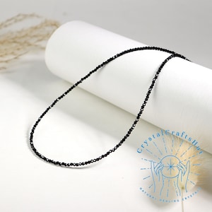 K&ouml;nnte beinhalten: Eine schwarze Perlenkette aus kleinen, facettierten schwarzen Onyxperlen. Die Kette liegt auf einer wei&szlig;en Oberfl&auml;che.