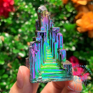 Peut inclure: Un grand cristal de bismuth iridescent, multi-facettes, vert, bleu et violet en forme de tour. Le cristal est tenu dans une main.