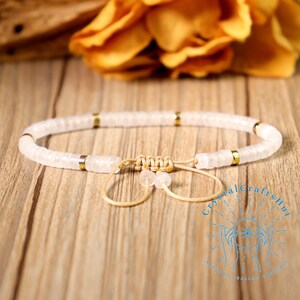 May include: White beaded bracelet with gold-colored accents. The bracelet features small, round beads strung together with a beige cord and a sliding knot closure. The bracelet is displayed on a wooden surface.