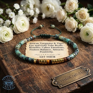 Op de afbeelding: Een turquoise en goudkleurige kralenarmband met de tekst "African Turquoise & Tiger Eye and Gold Tube Beads - Benefits: Calms Emotions, Fosters Balance, Attracts Positivity." De armband ligt op een houten oppervlak met witte bloemen.