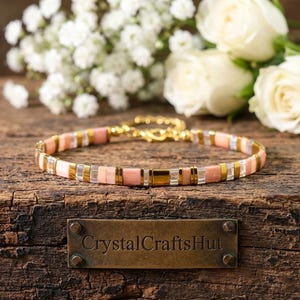 May include: A delicate bracelet featuring a pattern of pink, gold, and clear beads. The bracelet is set against a rustic wooden surface, with white flowers in the background. The bracelet has a gold-colored clasp.