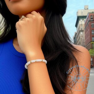 May include: A white bracelet with gold-colored accents is worn on a wrist. The bracelet is made of round, white beads separated by small gold-colored cylindrical beads. The person is wearing a blue top.