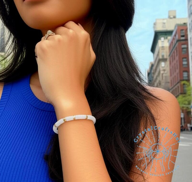 May include: A white bracelet with gold-colored accents is worn on a wrist. The bracelet is made of round, white beads separated by small gold-colored cylindrical beads. The person is wearing a blue top.