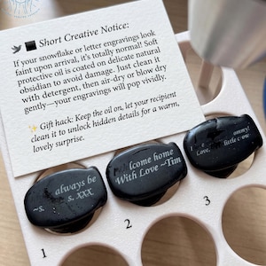 May include: Three black obsidian stones with engraved messages, including "always be s. XXX", "Welcome home With Love ~Tim", and "I love ommy! little crow". The stones are in a light pink holder with a card that has text.