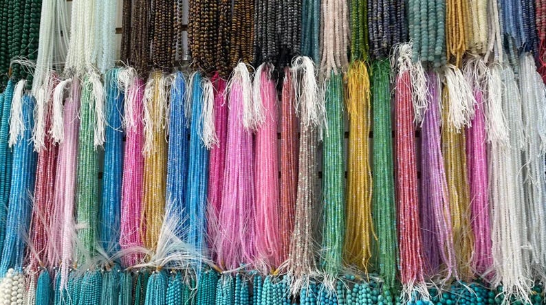 Pu&ograve; includere: Una variet&agrave; di fili di perline colorate appese a un muro. I fili sono fatti di diversi materiali, tra cui vetro, metallo e legno. Le perline sono di varie forme e dimensioni, e i colori sono vivaci e vibranti.
