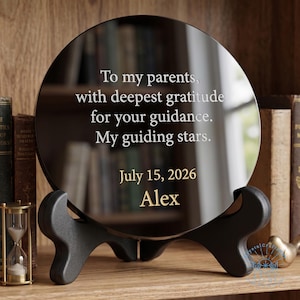 May include: A circular black reflective plaque held by a black stand. The text reads: To my parents, with deepest gratitude for your guidance. My guiding stars. July 15, 2026. Alex. The plaque features a smooth, glossy surface with engraved lettering.