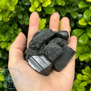 以下が含まれることがあります： 手の中に黒いトルマリンの結晶の山が置かれています。結晶は粗く、自然な研磨されていない外観をしています。濃い黒色で、銀色の筋がいくつか入っています。
