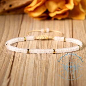 May include: A white beaded bracelet with gold accents. The bracelet features small, flat, round beads strung together with gold-colored spacers. The adjustable cord is a light beige color. The background is a wooden surface with a blurred yellow flower.