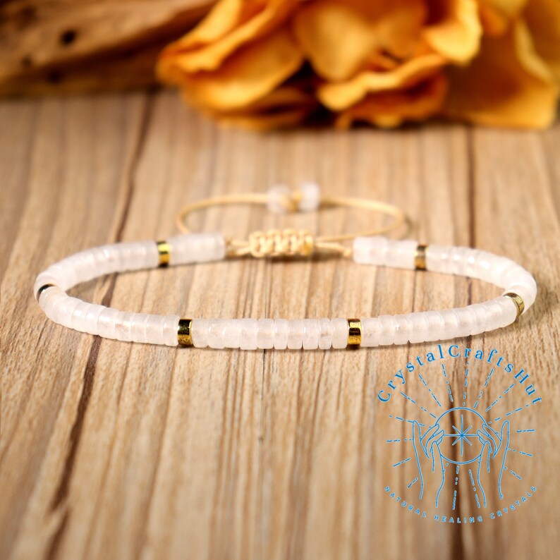 May include: A white beaded bracelet with gold accents. The bracelet features small, flat, round beads strung together with gold-colored spacers. The adjustable cord is a light beige color. The background is a wooden surface with a blurred yellow flower.