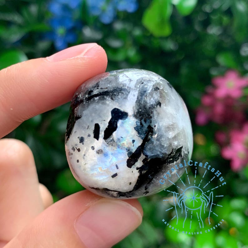 May include: A smooth, round, white and black gemstone with iridescent rainbow colors. The stone is being held in a hand.