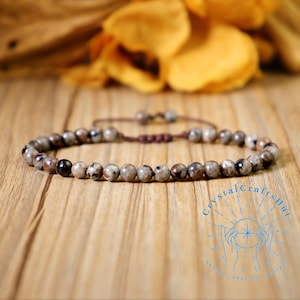 May include: A brown and gray beaded bracelet with a sliding knot closure. The bracelet is made of natural stones and is on a wooden surface. The image includes the text "Crystal Crafts Hut Natural Healing Crystals".