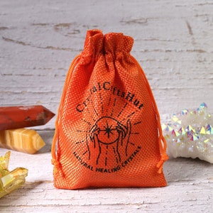 May include: An orange drawstring bag with the text "Crystal Gifts Hut" and "Natural Healing Crystals" printed in black. The bag is made of a textured material and is filled with crystals. Other crystals are visible in the background.